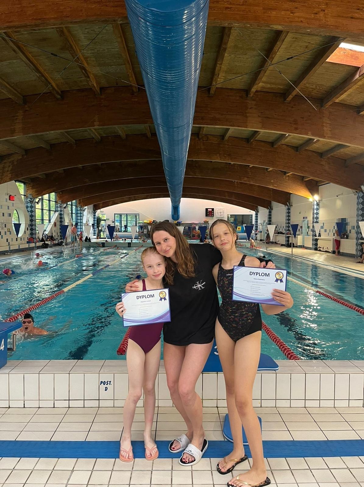 Instruktorka pływania z dwiema uczennicami w strojach kąpielowych, trzymające dyplomy ukończenia kursu na tle basenu krytego z osobami pływającymi.