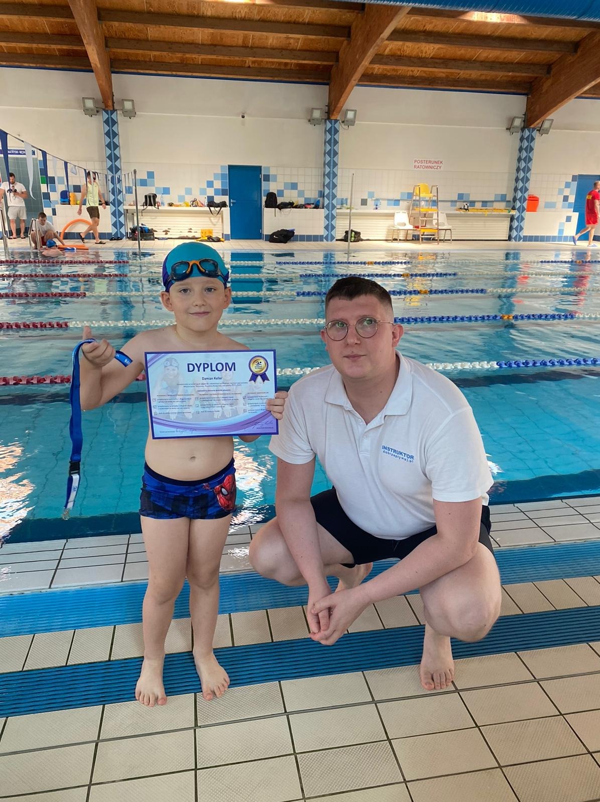 Chłopiec w stroju kąpielowym i czepku pływackim trzyma dyplom i medal, obok kuca instruktor na tle basenu.