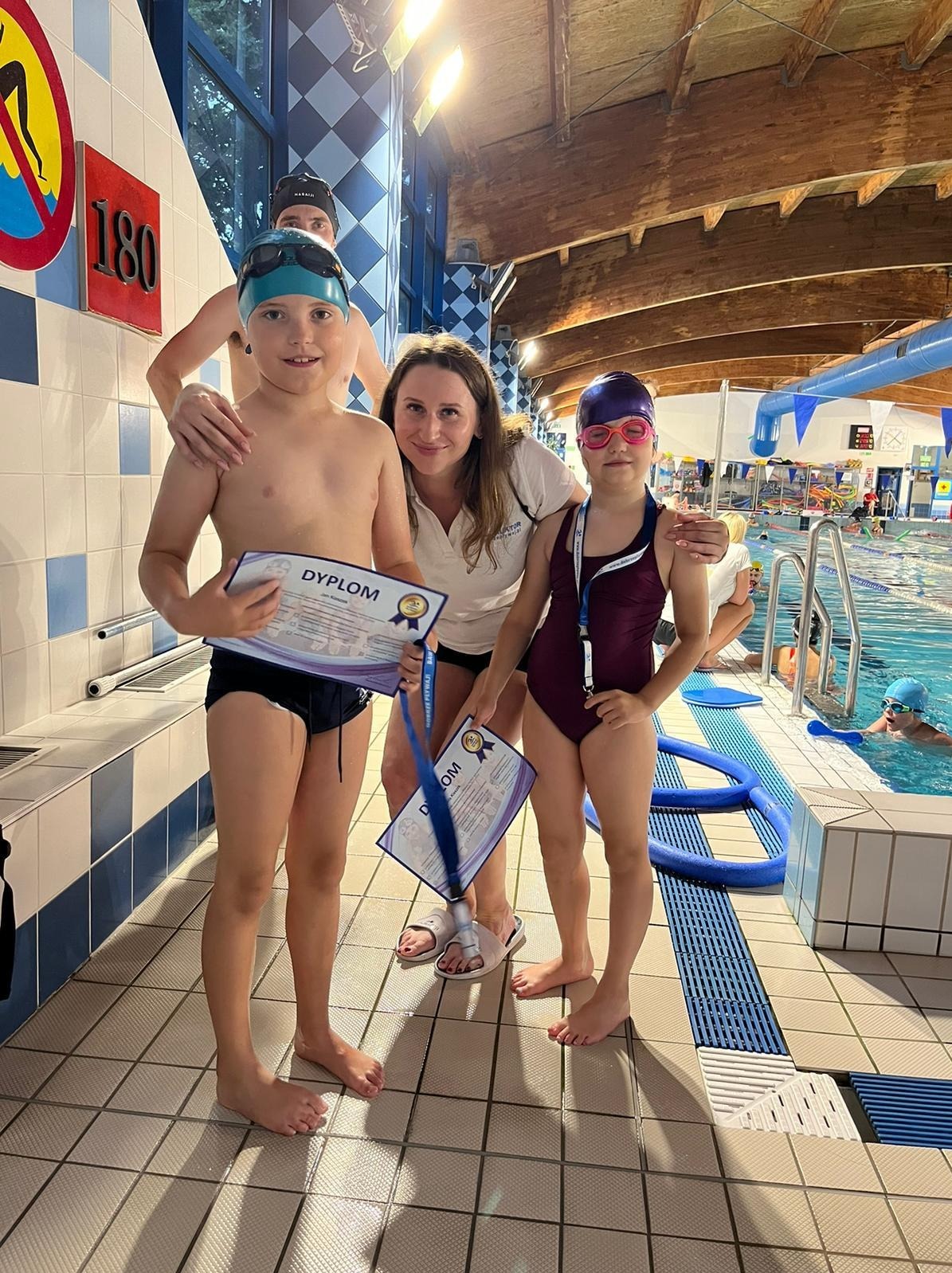 Dwoje dzieci z dyplomami po lekcjach pływania, obok instruktorka na tle basenu i osoby w tle
