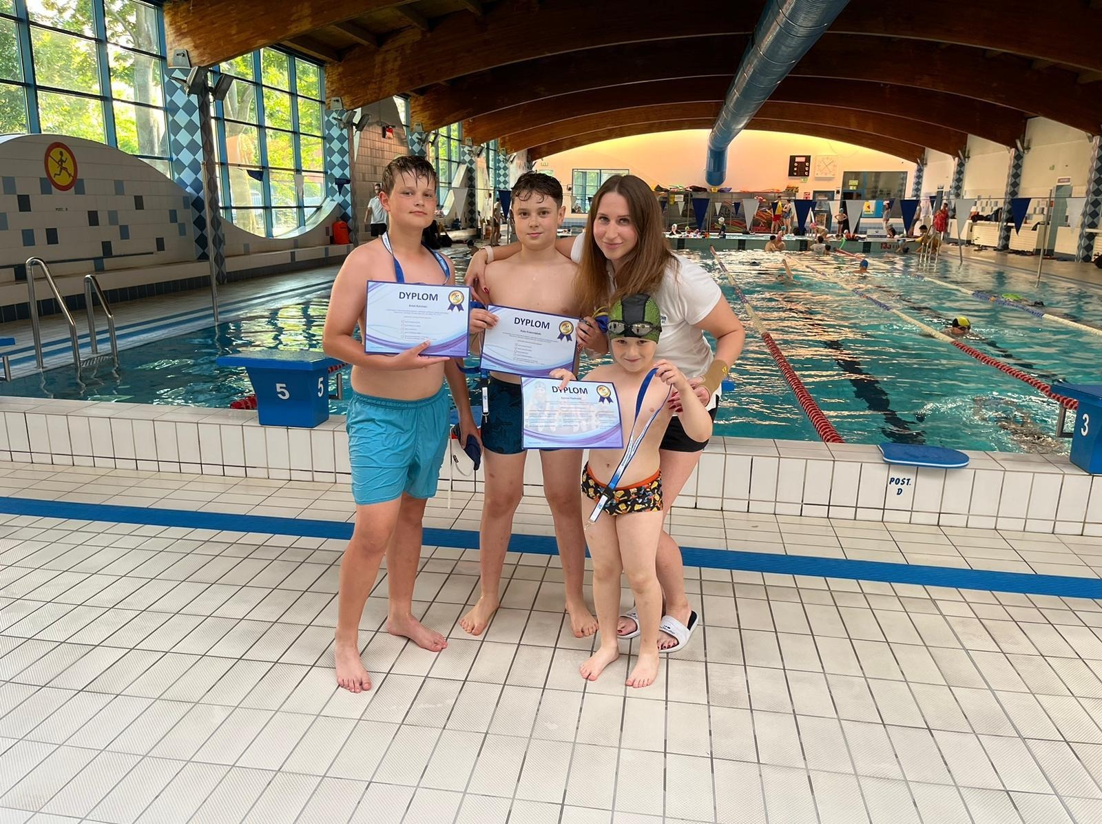 Trójka chłopców w strojach kąpielowych i kobieta stoją na tle basenu, trzymając dyplomy ukończenia kursu pływania.