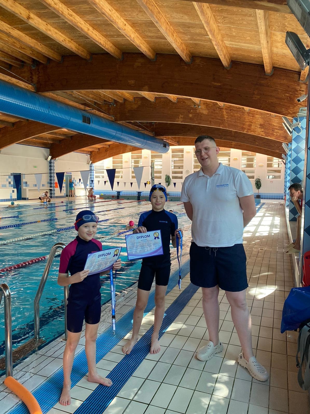 Dwoje dzieci w strojach pływackich, z dyplomami i medalami, pozuje z instruktorem na tle krytego basenu.