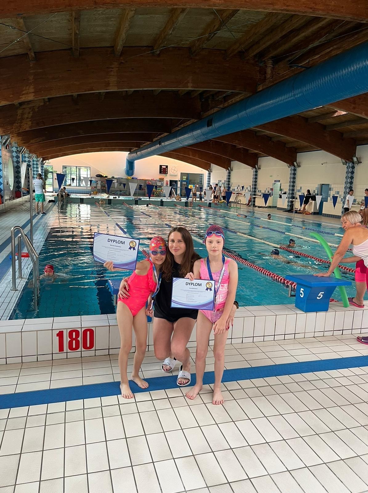 Dwie dziewczynki w strojach kąpielowych z dyplomami i trenerka pozują na tle basenu krytego z pływającymi osobami.