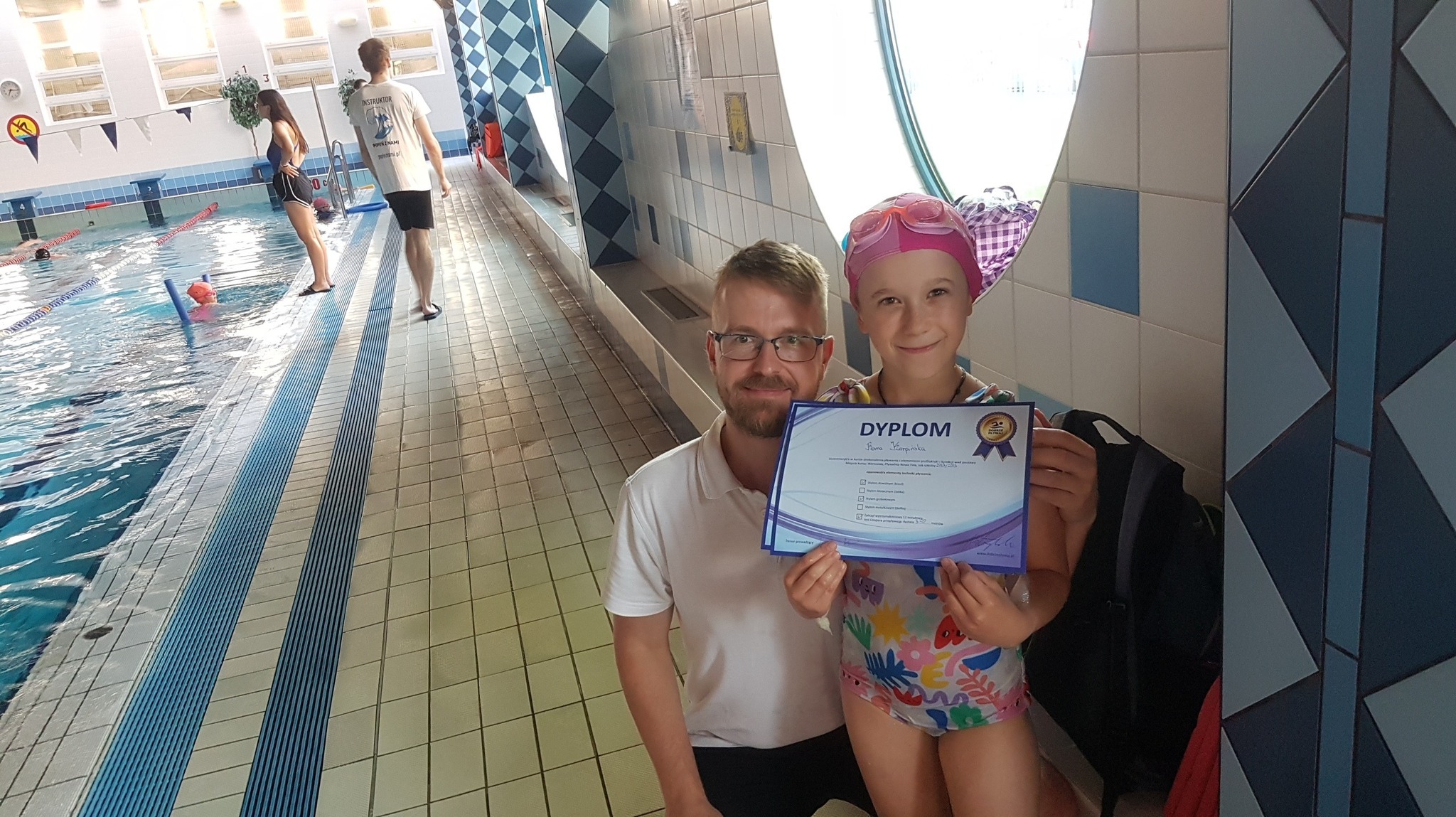 Instruktor pływania wręcza dyplom dziewczynce w czepku na tle basenu z innymi osobami.