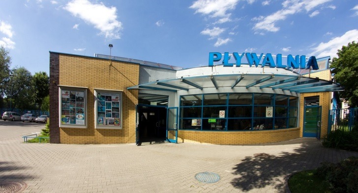 Fasada budynku pływalni z niebieskim napisem 'PŁYWALNIA' na dachu, widoczne plakaty reklamowe w oknach i parking z zaparkowanymi samochodami.