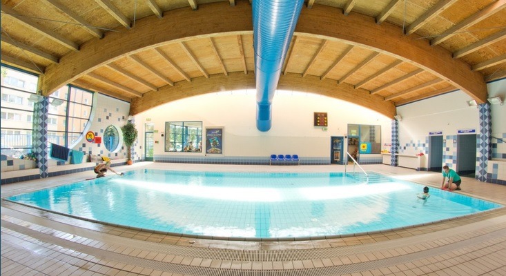 Panoramiczne ujęcie krytego basenu z jasną wodą, drewnianą konstrukcją dachu i osobami w wodzie; w tle widoczne okna i dekoracje ścienne.