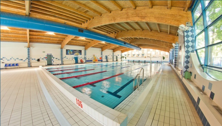 Kryty basen sportowy z wyznaczonymi torami pływackimi, oświetlony drewnianą konstrukcją dachu i niebieskimi rurami wentylacyjnymi w Warszawie.