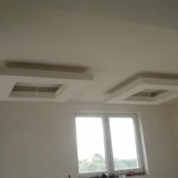 Wykończone wnętrze z tynkami gipsowymi i cementowo-wapiennymi, widoczne dwa kwadratowe podwieszane sufity z otworami na oświetlenie, ściany pomalowane na jasny kolor, okno z widokiem na zieleń.