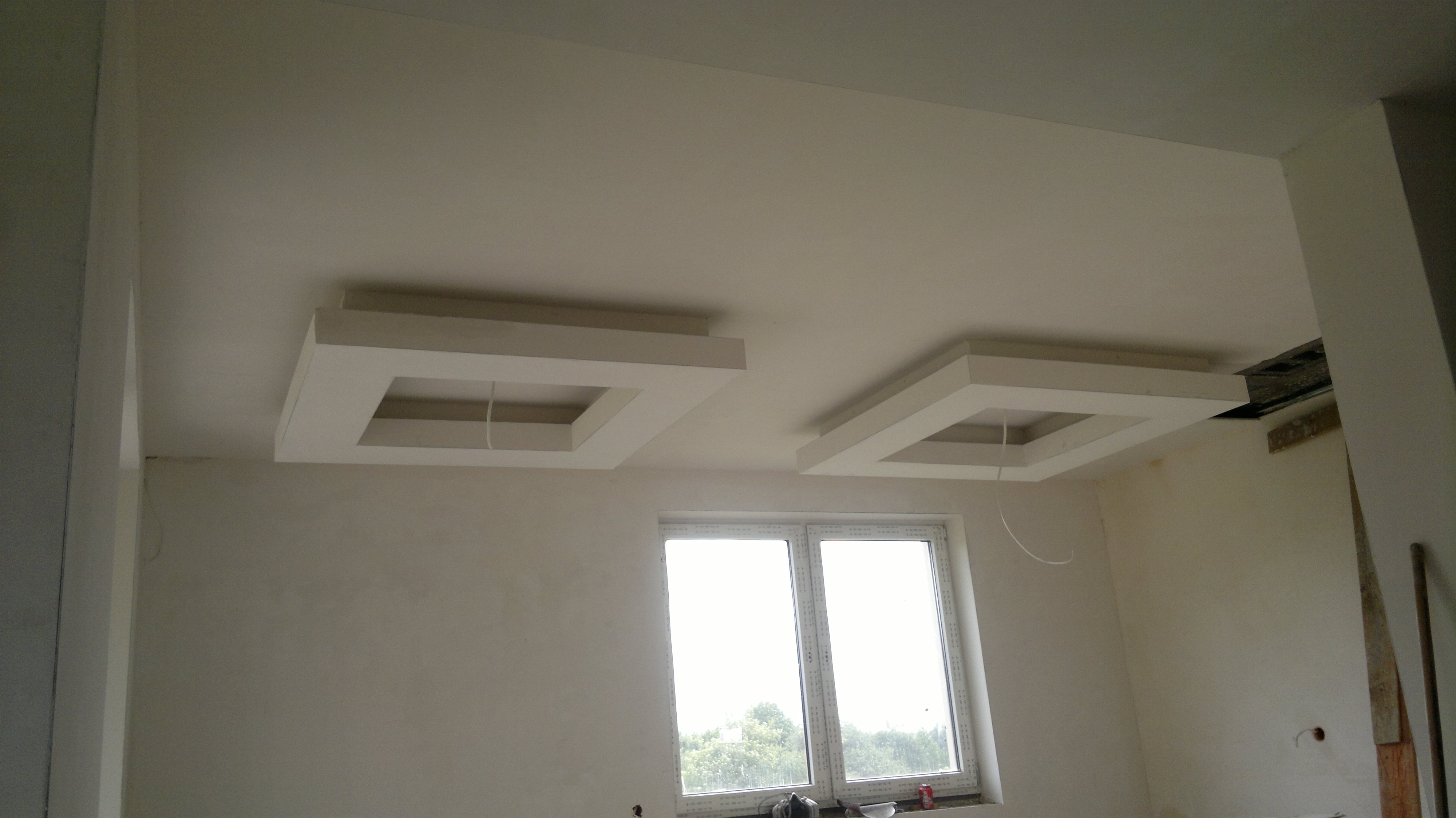 Wykończone wnętrze z tynkami gipsowymi i cementowo-wapiennymi, widoczne dwa kwadratowe podwieszane sufity z otworami na oświetlenie, ściany pomalowane na jasny kolor, okno z widokiem na zieleń.