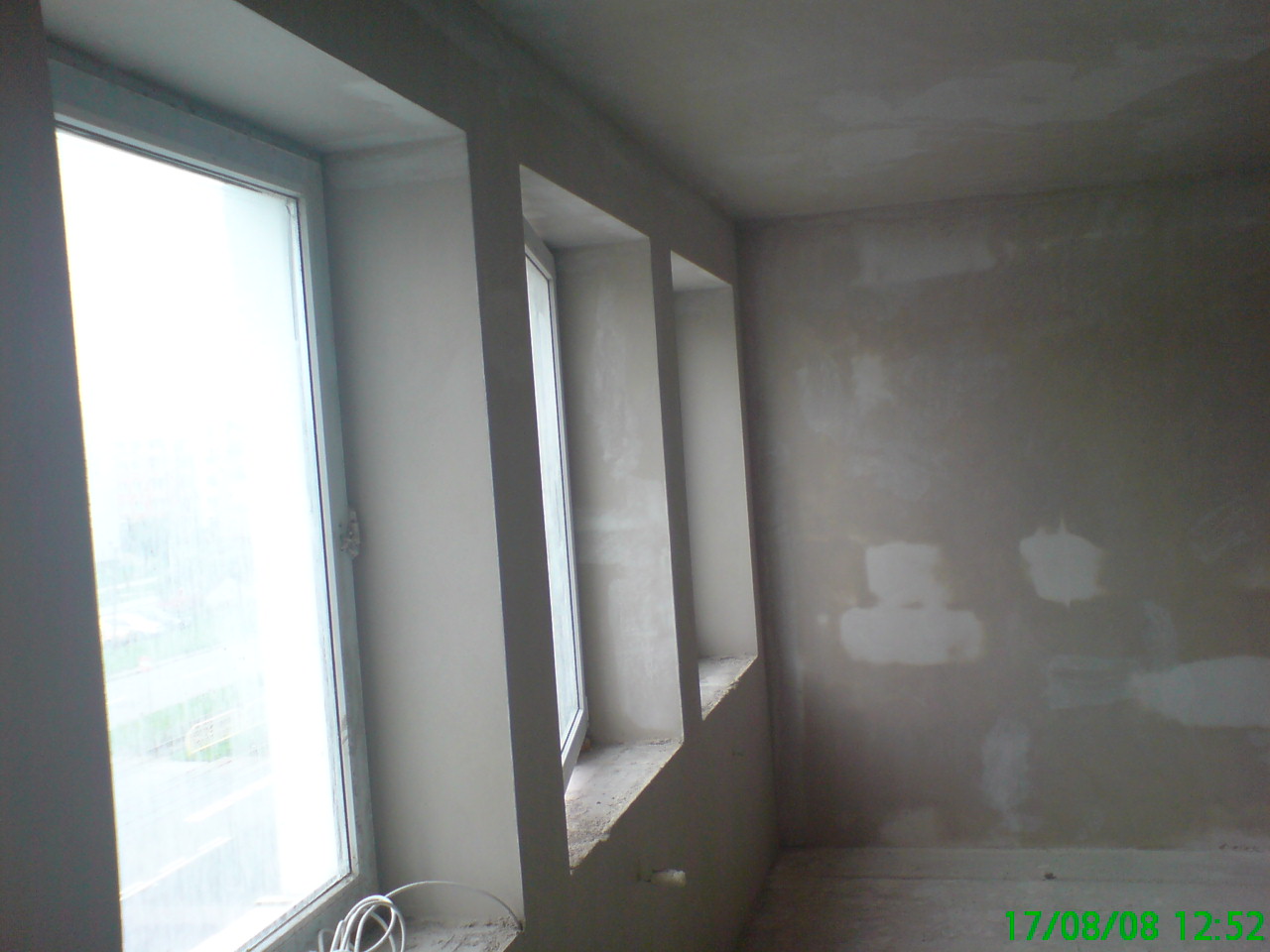 Surowa ściana z widocznymi łatami gipsowymi przed malowaniem, trzy okna w głębi z widokiem na zewnątrz, przewody elektryczne zwisające z parapetu.