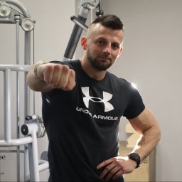 Konrad Janiszewski - Mężczyzna w czarnej koszulce Under Armour z zaciśniętą pięścią, pozujący na tle maszyny do ćwiczeń w siłowni.