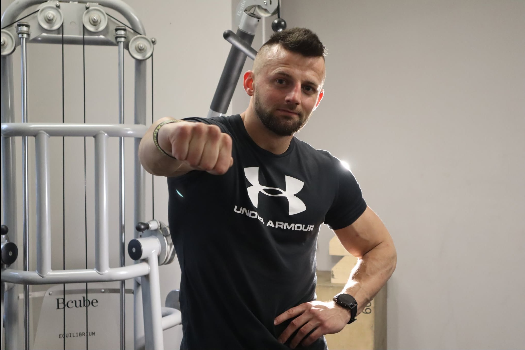 Mężczyzna w czarnej koszulce Under Armour z zaciśniętą pięścią, pozujący na tle maszyny do ćwiczeń w siłowni.