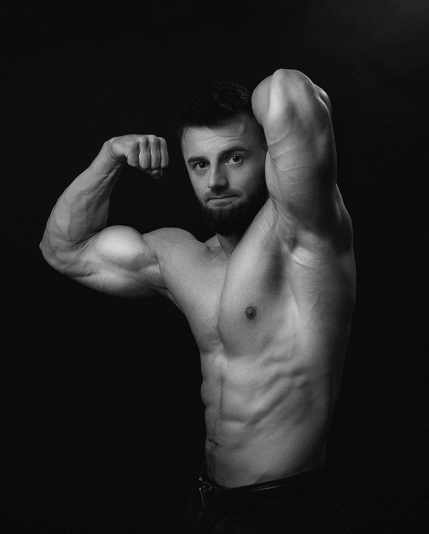 Czarno-białe zdjęcie mężczyzny z brodą, prezentującego muskulaturę bicepsów na ciemnym tle.