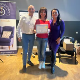 Trzy osoby pozują z certyfikatem w pomieszczeniu z banerem reklamowym i flipchartem. Kobieta w czerwonej bluzce trzyma certyfikat. Za nią stoi mężczyzna w białej koszuli i kobieta w fioletowej...