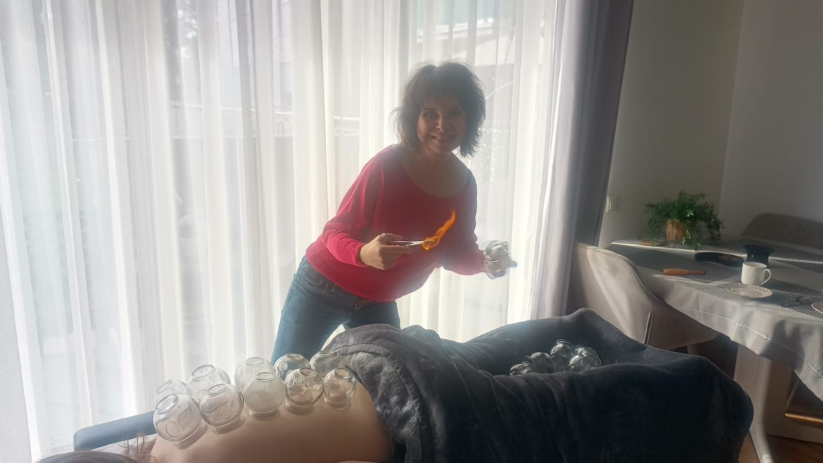 Kobieta w czerwonej bluzce, z płomieniem w szczypcach, przygotowuje bańki ogniowe na plecach pacjenta leżącego na stole przykrytym szarym kocem, z widocznymi bańkami już nałożonymi.