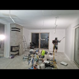 JAN-BUD - Malowanie ścian w trakcie remontu mieszkania, widoczne narzędzia malarskie, drabina, oraz wykończony kominek z jasnego kamienia.