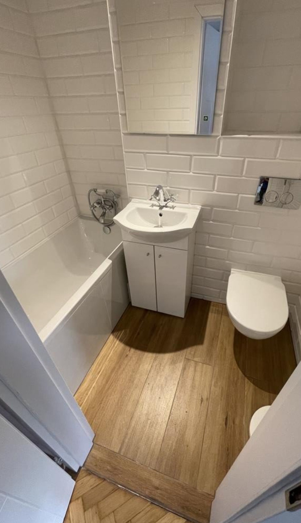 Wnętrze wykończonej łazienki z białą armaturą, glazurą w stylu cegiełek i drewnopodobną podłogą, widok z perspektywy wejścia.