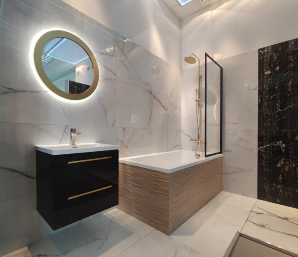 Nowoczesna łazienka z marmurowymi płytkami, okrągłym lustrem z podświetleniem LED, czarną szafką pod umywalką z złotymi uchwytami i wanną z drewnopodobną obudową, złota armatura.