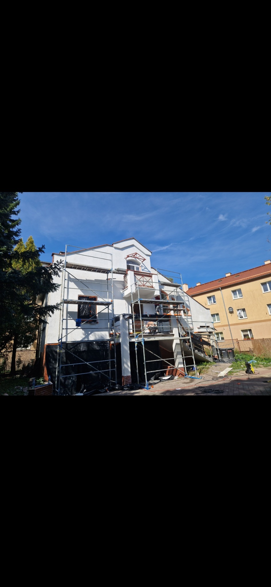 Dwupiętrowy dom w trakcie renowacji, otoczony rusztowaniem, zabezpieczony folią, widoczny fragment ogrodu i sąsiedni budynek.