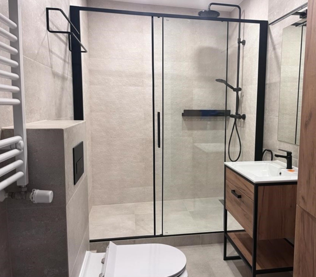 Nowoczesna łazienka z kabiną prysznicową typu walk-in z czarnymi profilami, umywalką na szafce z drewnianym frontem i czarną armaturą, grzejnikiem drabinkowym oraz podtynkowym systemem WC.