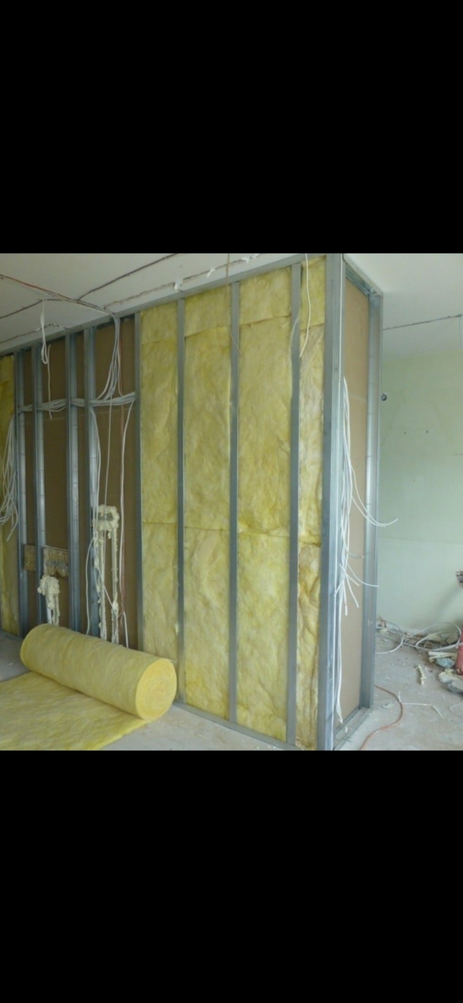 Montaż izolacji z wełny mineralnej w metalowym stelażu, widoczne przewody elektryczne i rolka materiału izolacyjnego na podłodze.