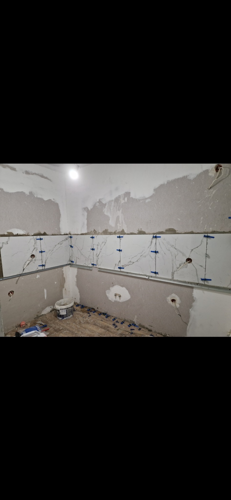Montaż płytek imitujących marmur na ścianie w kuchni, widoczne niezagruntowane fragmenty ściany, poziomica i niebieskie krzyżyki dystansowe, na podłodze resztki zaprawy i wiadro po farbie.