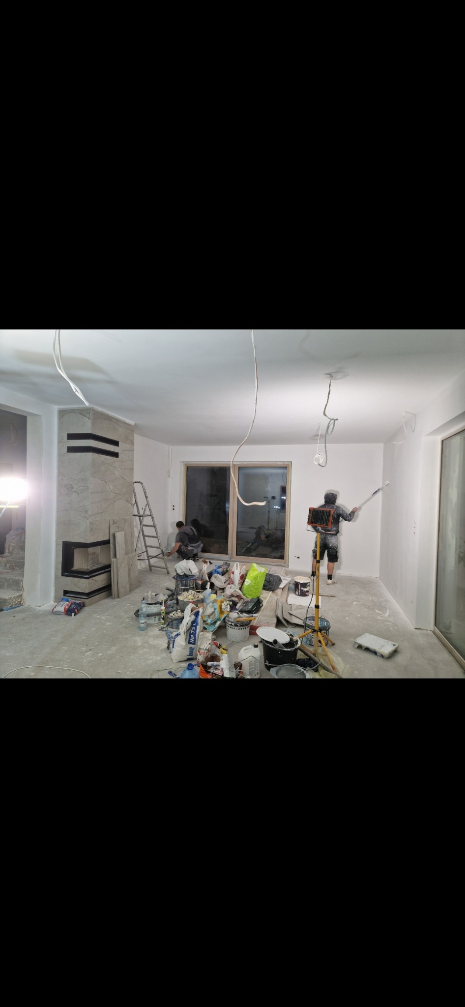 Malowanie ścian w trakcie remontu mieszkania, widoczne narzędzia malarskie, drabina, oraz wykończony kominek z jasnego kamienia.