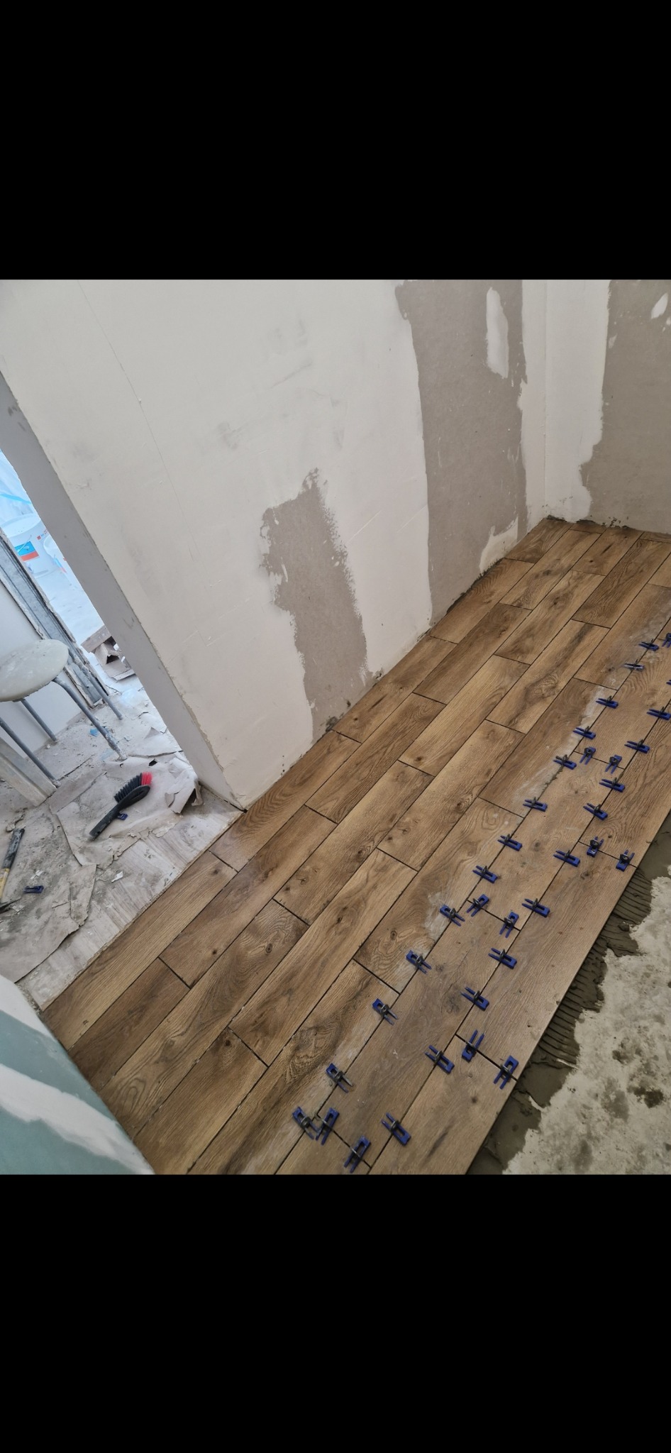 Układanie drewnopodobnych płytek podłogowych w trakcie prac, z użyciem niebieskich krzyżyków dystansowych, przy częściowo wykończonych ścianach.