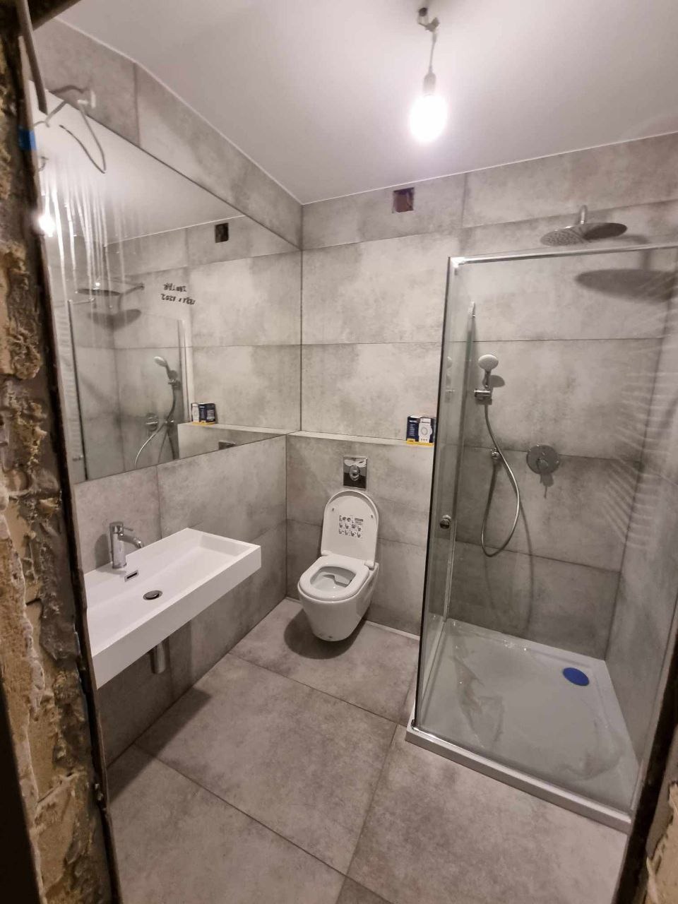 Nowoczesna łazienka z szarą płytką, wiszącą umywalką i kabiną prysznicową typu walk-in z niebieskim akcentem na brodziku.