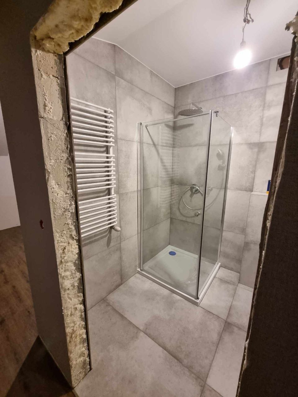 Wykończona łazienka z szarą glazurą, kabiną prysznicową z brodzikiem i grzejnikiem drabinkowym; surowy fragment ściany z pianką montażową.