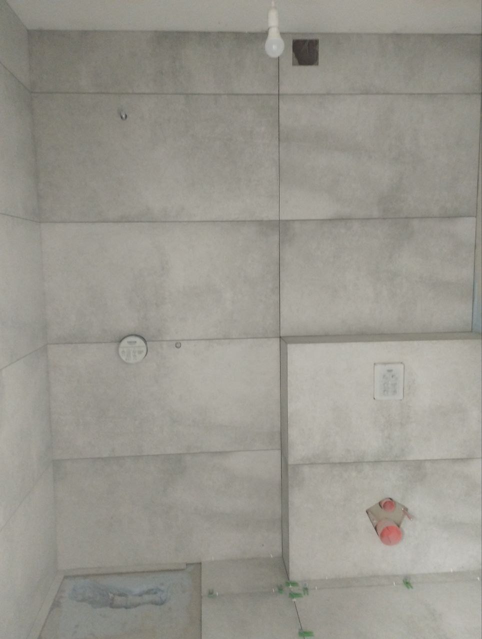 Ściana w trakcie kładzenia płytek w łazience, widoczne krzyżyki dystansowe, surowa żarówka i otwór instalacyjny.