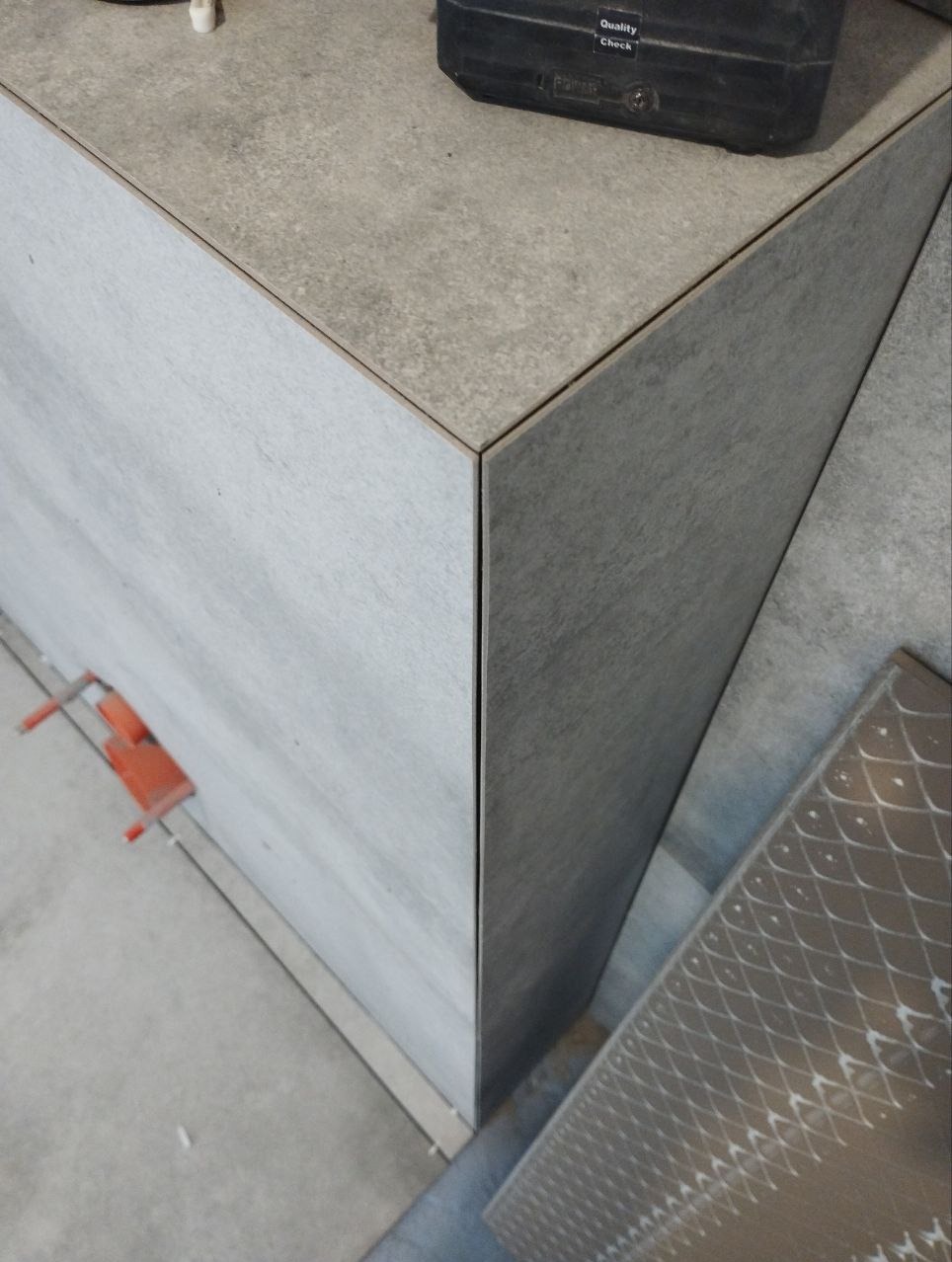 Wykończony narożnik obudowy z szarych płytek imitujących drewno, z pomarańczowym klipsem poziomującym i czarną skrzynką narzędziową w tle.