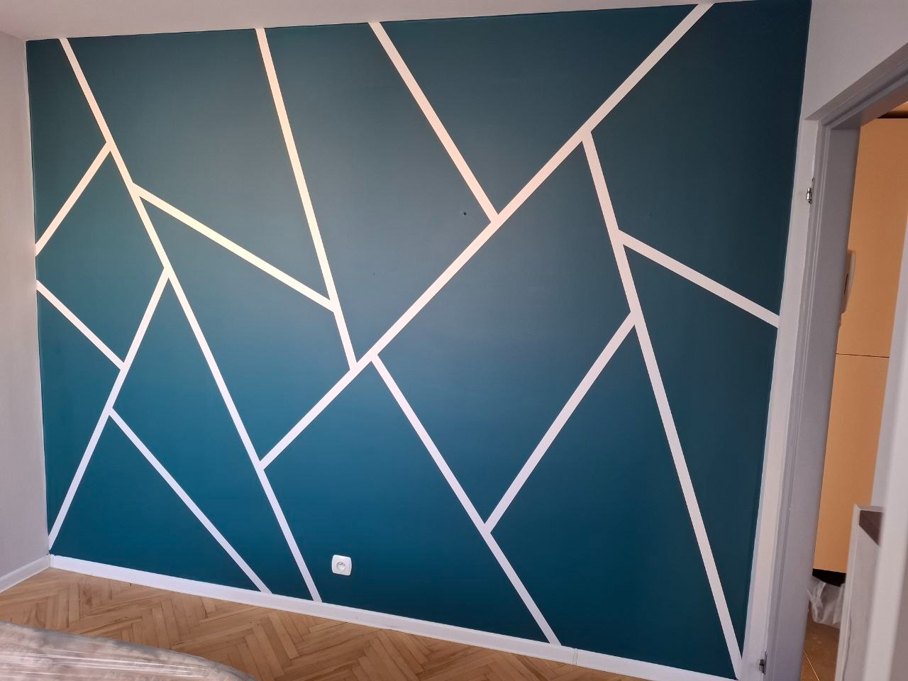 Ściana w kolorze morskiej zieleni z geometrycznym wzorem z białych linii w mieszkaniu w Olsztynie, widoczny parkiet i fragment otwartych drzwi.