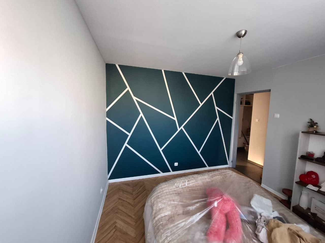 Ściana pomalowana na granatowo z białymi geometrycznymi wzorami, widoczny fragment parkietu i łóżka przykrytego folią ochronną, lampa sufitowa.