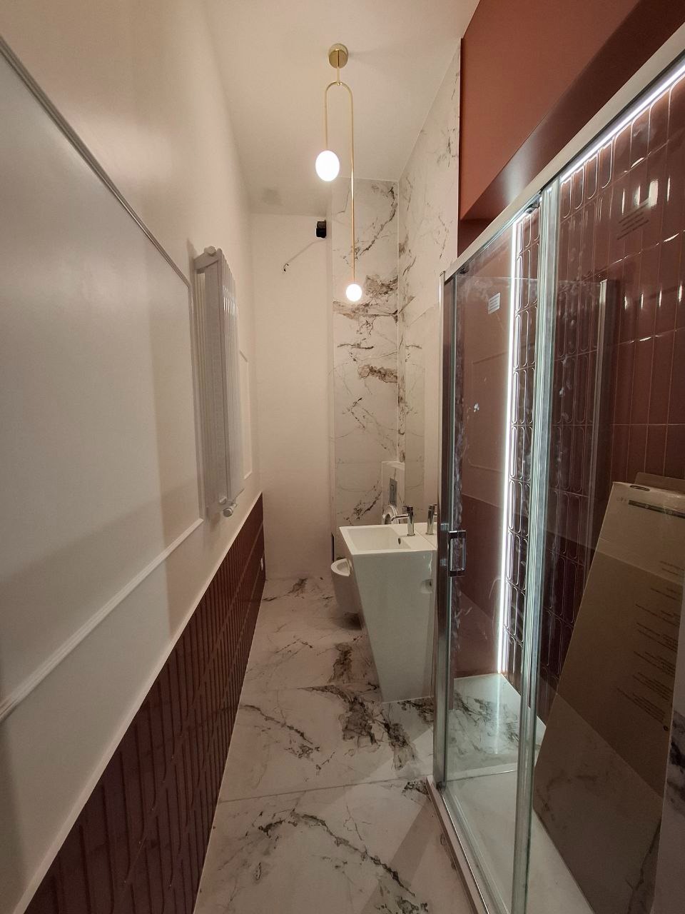 Wąska łazienka po remoncie z marmurową podłogą, wiszącą umywalką na postumencie, kabiną prysznicową z przesuwnymi drzwiami i dekoracyjnym oświetleniem w postaci dwóch kulistych lamp na złotym stelażu.