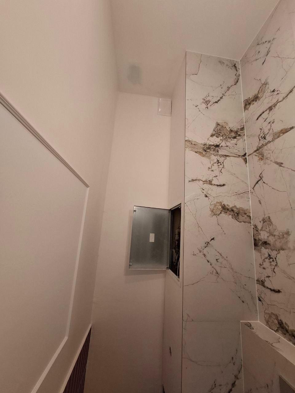 Wnętrze łazienki po remoncie, z płytkami imitującymi marmur na ścianach, otwartą metalową szafką z instalacją elektryczną i widoczną wentylacją.