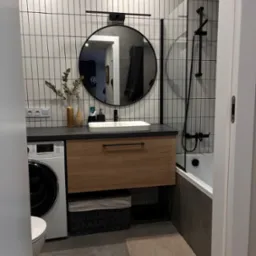 Nowoczesna łazienka z umywalką nablatową, drewnianą szafką, pralką, okrągłym lustrem w czarnej ramie, białymi płytkami ściennymi z czarną fugą oraz wanną z czarną armaturą.