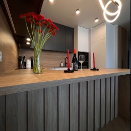 ARTIUM CRAFT SPÓŁKA Z OGRANICZONĄ ODPOWIEDZIALNOŚCIĄ - Nowoczesna aranżacja kuchni z wyspą, dekorowana czerwonymi gerberami w wazonie, butelką wina i świecami, oświetlona designerską lampą.
