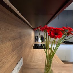 ARTIUM CRAFT SPÓŁKA Z OGRANICZONĄ ODPOWIEDZIALNOŚCIĄ - Jasny blat kuchenny z drewnianym wykończeniem, na którym stoi przezroczysty wazon z bukietem czerwonych gerber, w tle widoczna czarna płyta indukcyjna i białe szafki.