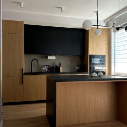 ARTIUM CRAFT SPÓŁKA Z OGRANICZONĄ ODPOWIEDZIALNOŚCIĄ - Nowoczesna kuchnia z drewnianymi frontami szafek, czarnymi elementami i wyspą kuchenną z blatem z czarnego kamienia, oświetlona lampą wiszącą z kulistymi kloszami.