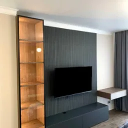 Nowoczesny salon z zabudową meblową w odcieniach szarości i drewna, obejmującą wysoką przeszkloną witrynę z oświetleniem, szafkę RTV pod telewizorem na tle ściany z pionowymi panelami oraz wiszący...