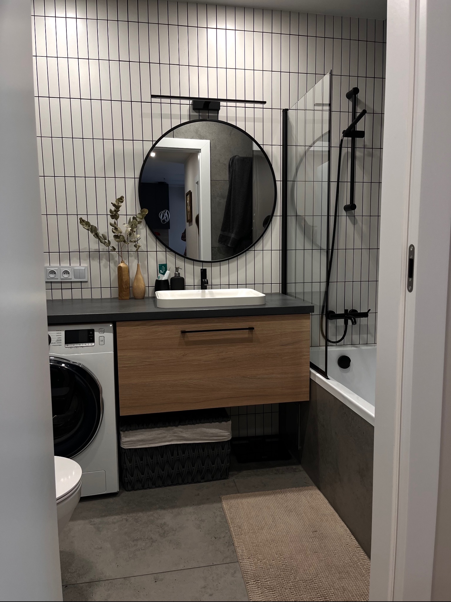 Nowoczesna łazienka z umywalką nablatową, drewnianą szafką, pralką, okrągłym lustrem w czarnej ramie, białymi płytkami ściennymi z czarną fugą oraz wanną z czarną armaturą.