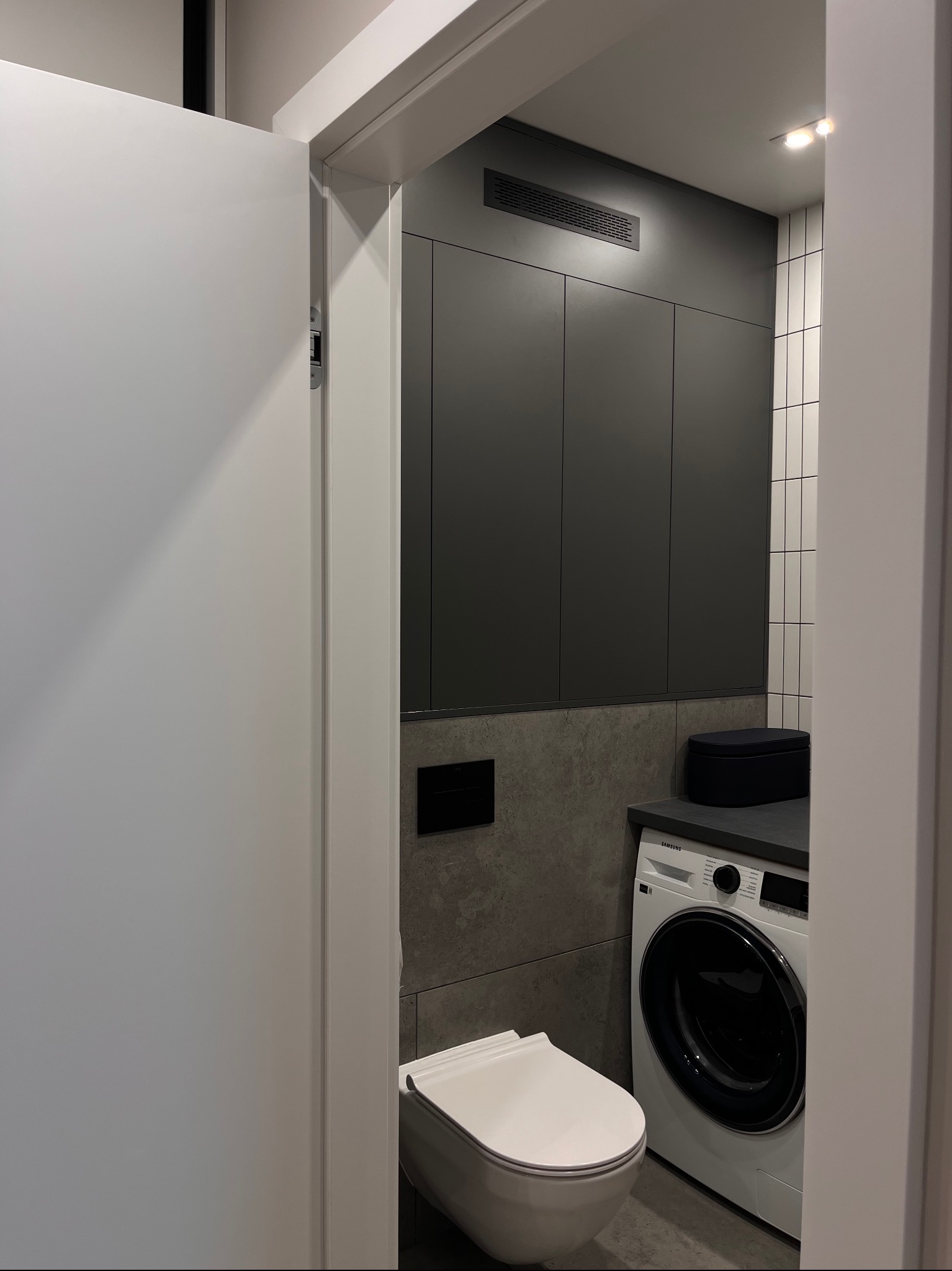 Nowoczesna łazienka z szarą zabudową meblową, pralką Samsung i podwieszaną toaletą, wykończona płytkami w odcieniach szarości i bieli, oświetlona punktowo.