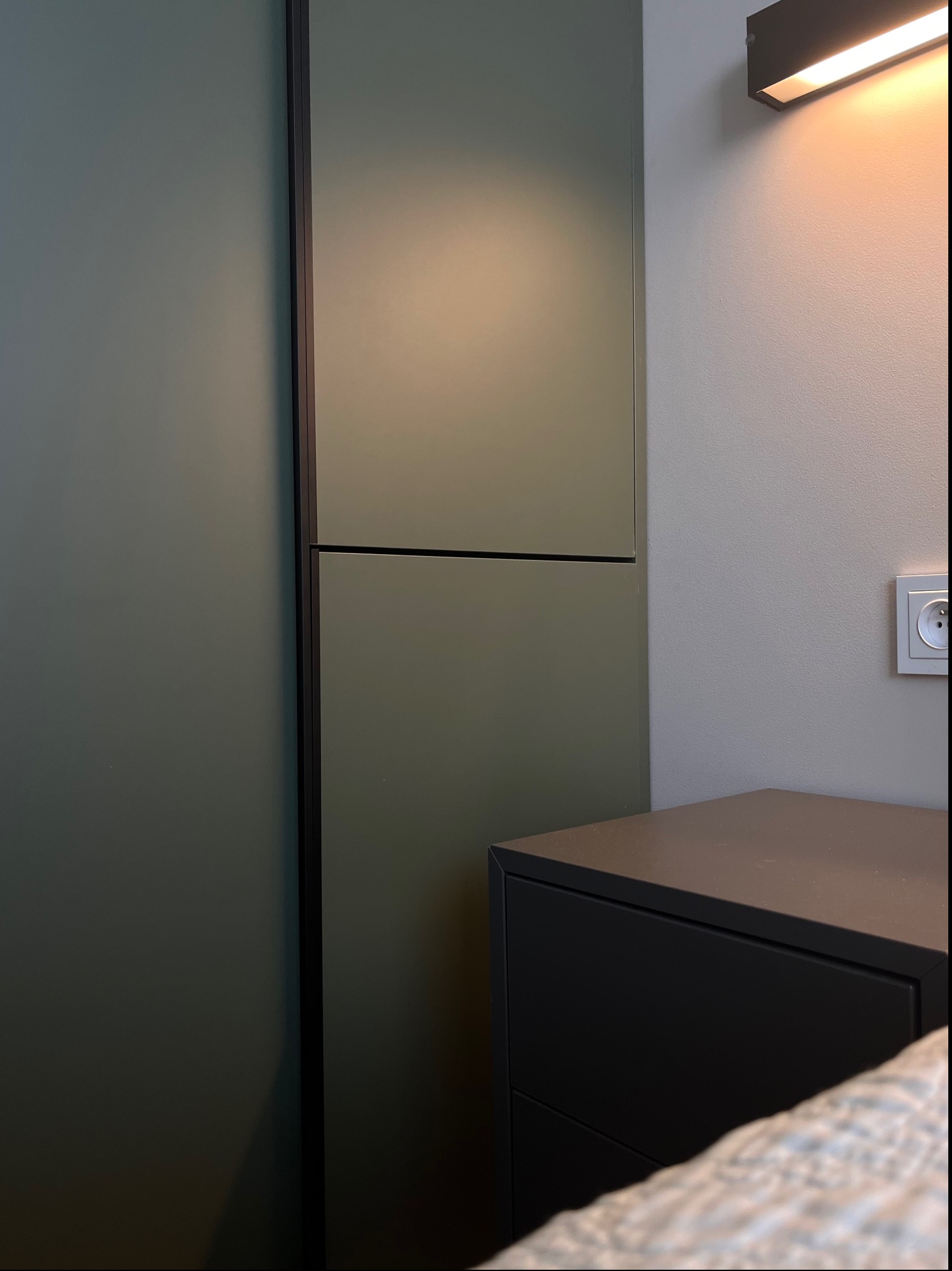 Fragment szafy wnękowej w odcieniach zieleni z czarnymi obramowaniami, obok szafka nocna i lampa ścienna, widoczny fragment pościeli.