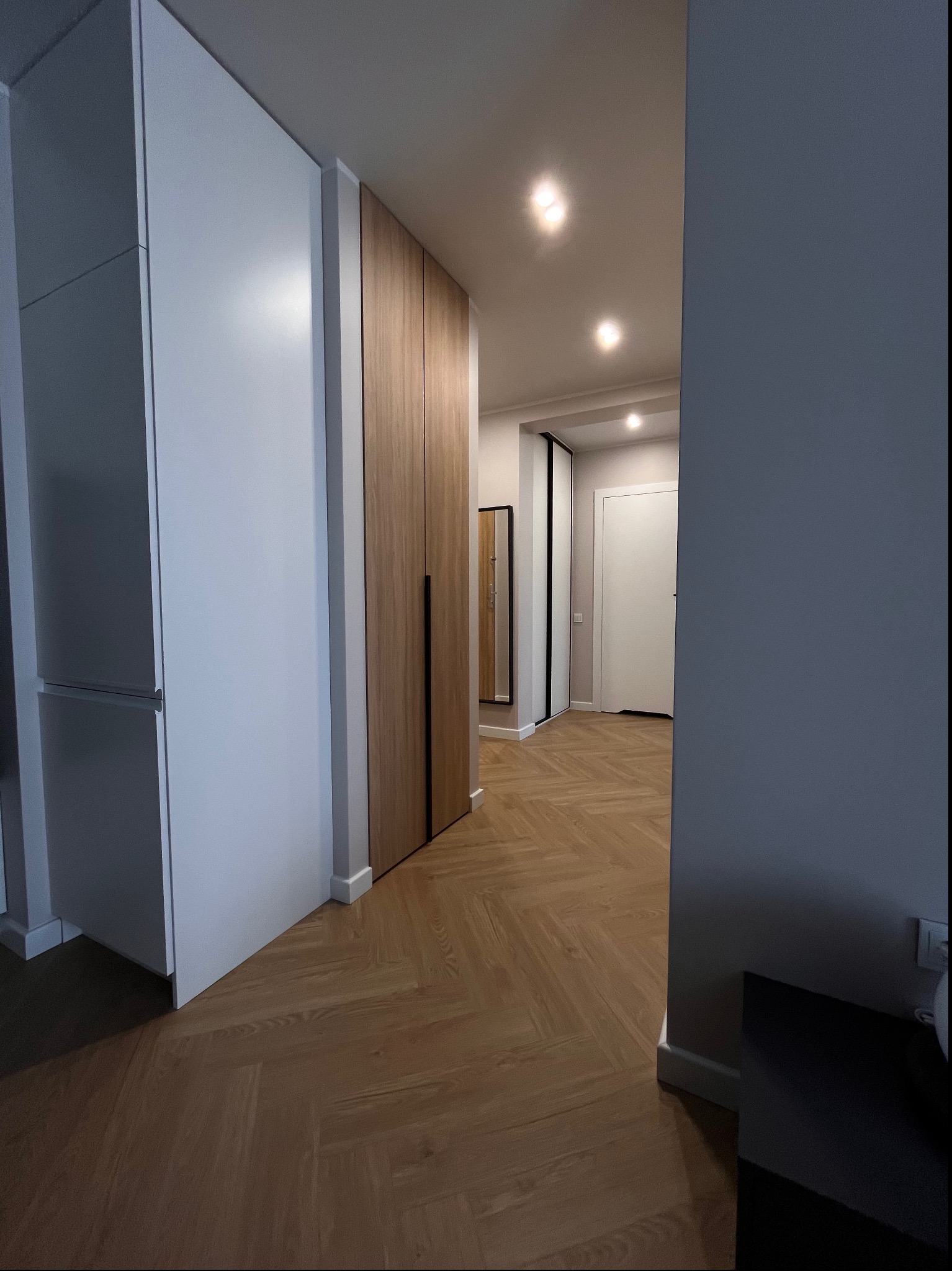 Wnętrze korytarza z zabudową szaf: biała szafa z szufladami i drewniana szafa z czarnymi uchwytami, parkiet w jodełkę, oświetlenie sufitowe punktowe.