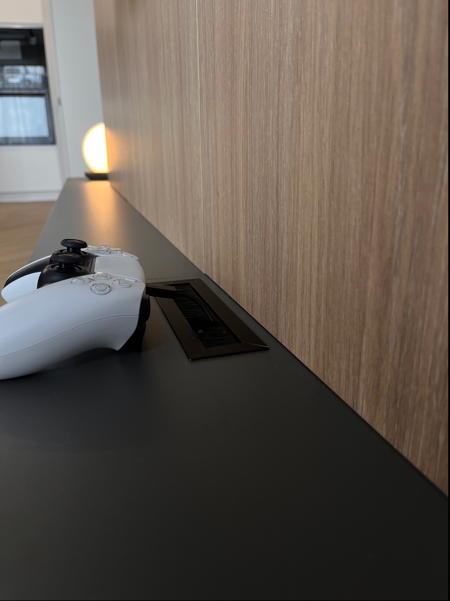 Nowoczesna szafka RTV z czarnym blatem i drewnianym panelem ściennym, na której leży biały kontroler do konsoli PlayStation 5 oraz widoczna jest czarna maskownica otworów na kable.