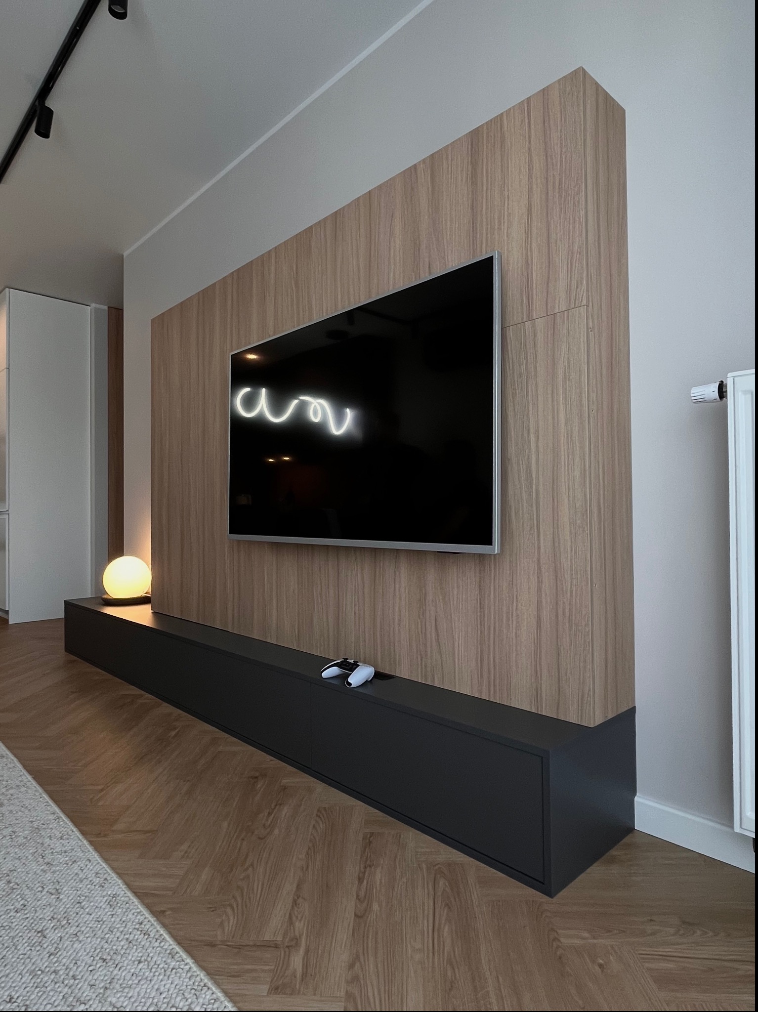Nowoczesny salon z drewnianą ścianą, na której wisi telewizor, pod nim czarna szafka RTV z kontrolerem do gier, a obok lampa kula