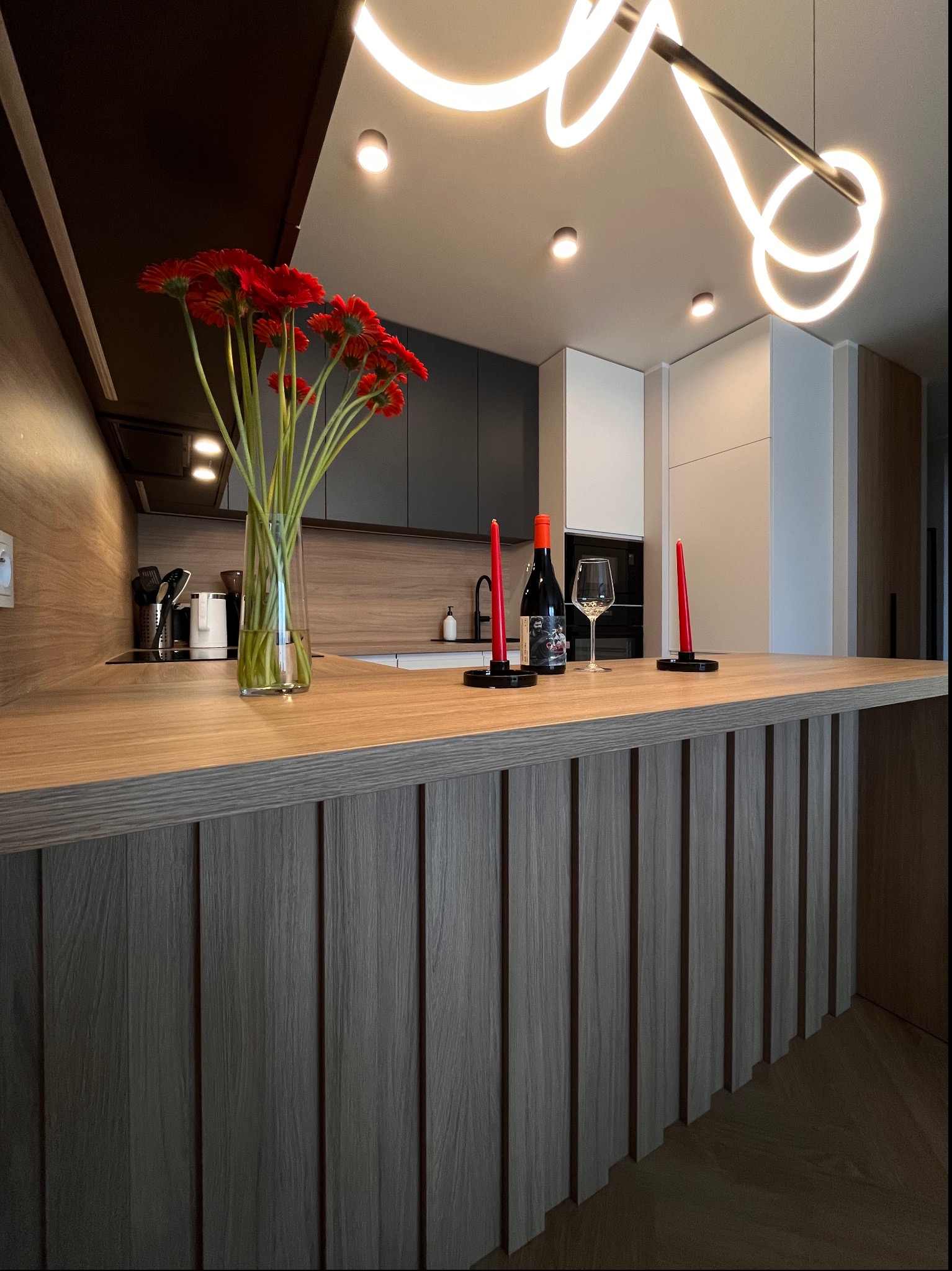 Nowoczesna aranżacja kuchni z wyspą, dekorowana czerwonymi gerberami w wazonie, butelką wina i świecami, oświetlona designerską lampą.