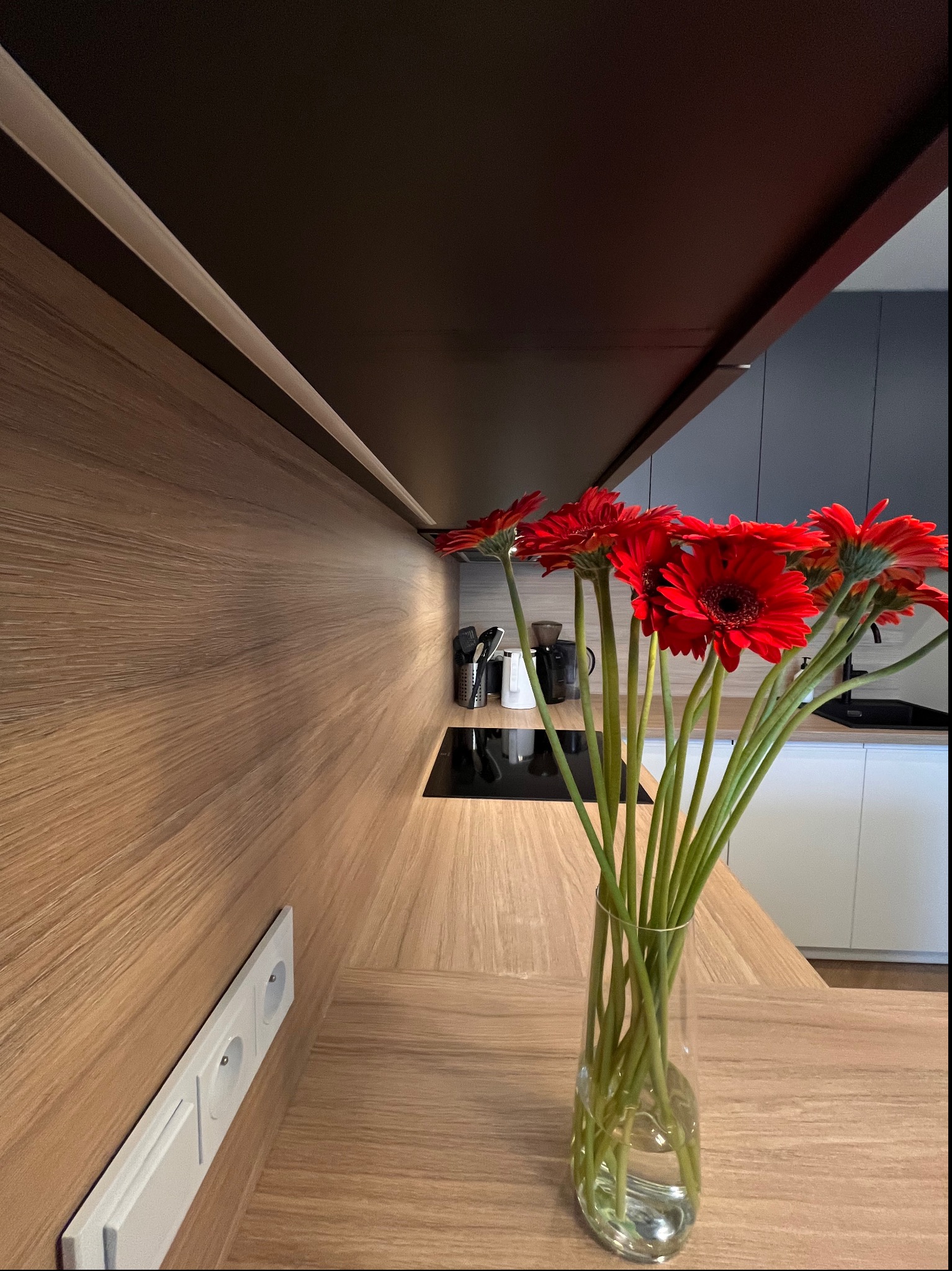Jasny blat kuchenny z drewnianym wykończeniem, na którym stoi przezroczysty wazon z bukietem czerwonych gerber, w tle widoczna czarna płyta indukcyjna i białe szafki.