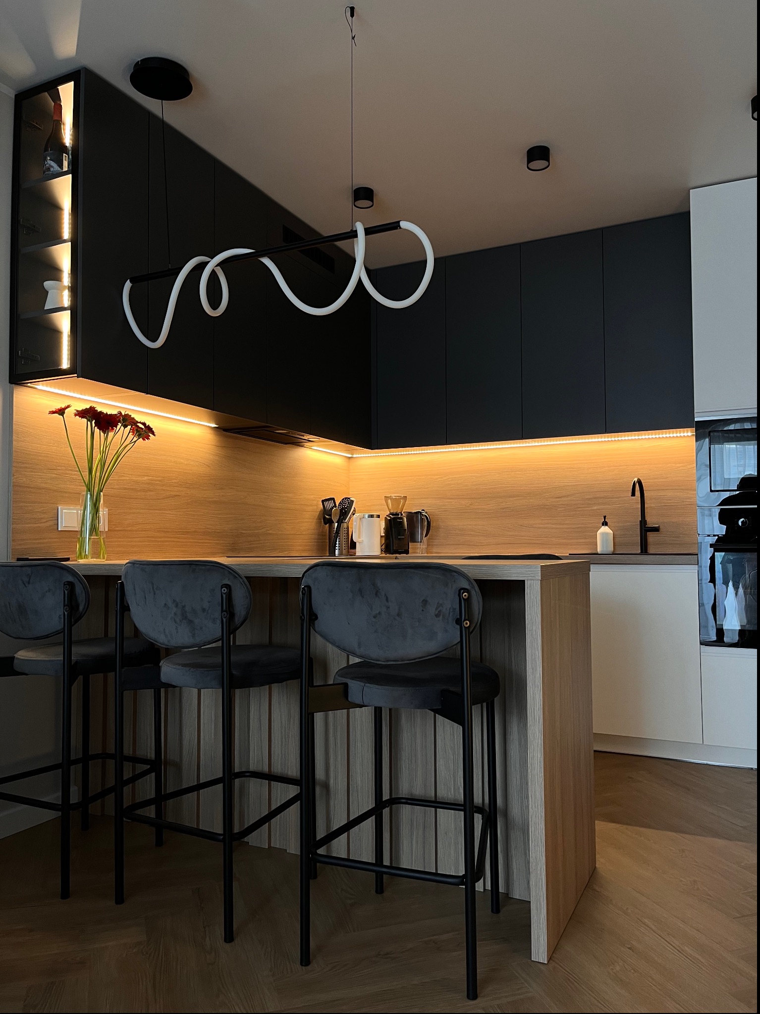 Nowoczesna kuchnia z czarnymi szafkami i drewnianym blatem, oświetlona listwą LED, z wyspą i trzema welurowymi hokerami na czarnych nogach, nad wyspą designerska lampa.