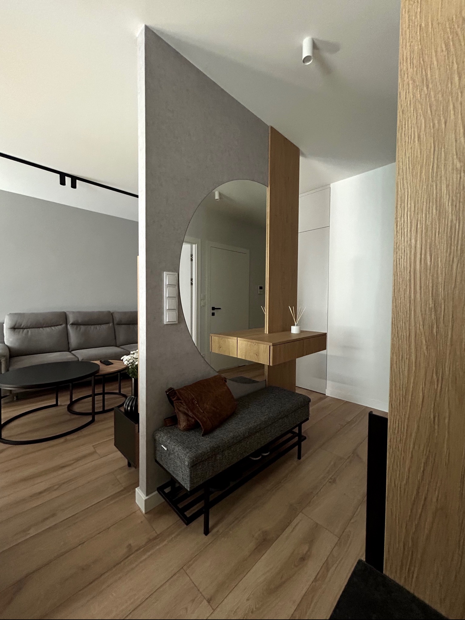 Nowoczesne wnętrze przedpokoju z siedziskiem, lustrem w okrągłej ramie i drewnianą konsolą, widoczne fragmenty salonu z szarą sofą.